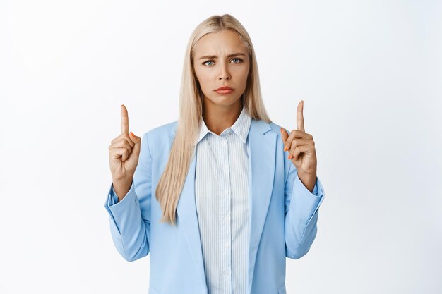 Femme d'affaires en colère pointant les doigts vers le haut en fronçant les sourcils et regardant avec déception debout en costume élégant sur fond blanc