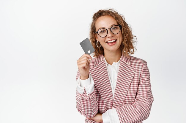 Femme d'affaires confiante montrant une carte de crédit et souriante, debout en costume et lunettes sur fond blanc