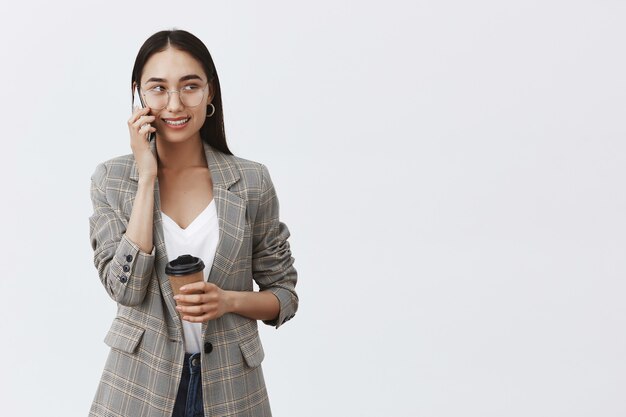 Femme d'affaires confiante en veste et lunettes, regardant à droite avec une expression intriguée et joyeuse tout en utilisant un smartphone et en buvant du café