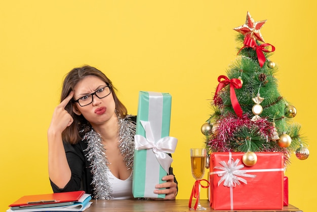 Femme d'affaires confuse en costume avec des lunettes montrant son cadeau et assis à une table avec un arbre de Noël dessus dans le bureau