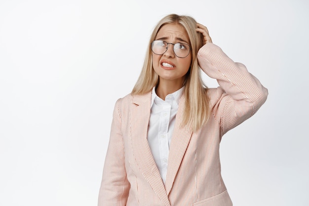 Une femme d'affaires confuse se gratte la tête et regarde avec un visage perplexe portant un costume et des lunettes sur fond blanc