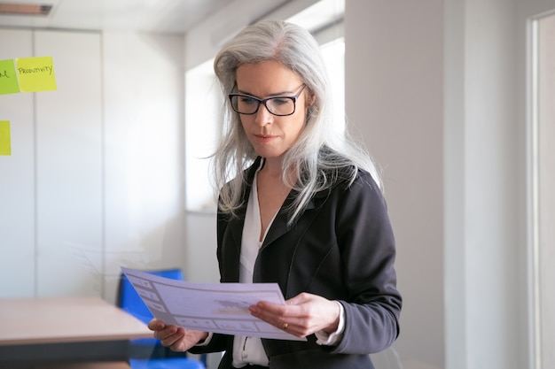 Femme d'affaires de contenu dans les statistiques de lecture de lunettes. Employeur aux cheveux gris concentré avec succès en costume debout dans la salle de bureau et tenant le document. Concept de marketing, d'affaires et de gestion