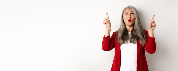 Photo gratuite femme d'affaires coréenne étonnée aux cheveux gris portant un blazer rouge au travail pointant les doigts vers le haut et regardant