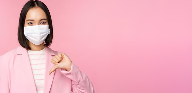 Femme d'affaires coréenne en masque médical et costume montre les pouces vers le bas n'aiment pas ou désapprouve le geste debout sur fond rose