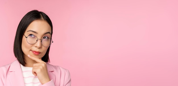 Femme d'affaires coréenne pensant porter des lunettes à la caméra réfléchie prenant une décision debout sur fond rose