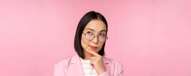 Femme d'affaires coréenne pensant porter des lunettes à la caméra réfléchie prenant une décision debout sur fond rose