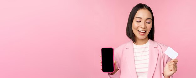 Femme d'affaires coréenne souriante en costume montrant la carte de crédit de l'écran du téléphone portable montrant l'interface de l'application bancaire en ligne fond rose