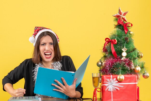 Femme d'affaires en costume avec chapeau de père Noël et décorations de nouvel an vérifiant le document se sentant nerveux et assis à une table avec un arbre de Noël dessus dans le bureau