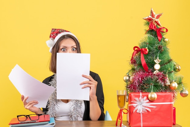 Femme D'affaires En Costume Avec Chapeau De Père Noël Et Décorations De Nouvel An Vérifiant Les Documents De Manière Surprenante Et Assis à Une Table Avec Un Arbre De Noël Dessus Dans Le Bureau