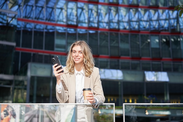 Femme d'affaires en costume debout avec smartphone dans la rue, boire du café à l'aide de l'application de téléphone mobile