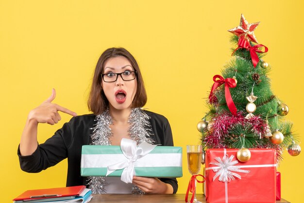 Femme d'affaires en costume avec des lunettes pointant son cadeau de manière surprenante et assis à une table avec un arbre de Noël dessus dans le bureau