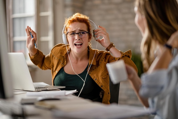 Femme d'affaires créative avec des écouteurs s'excusant de ne pas avoir entendu son collègue lui parler pendant qu'elle travaillait au bureau
