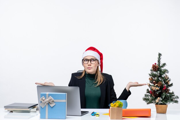 Femme d'affaires curieuse positive avec chapeau de père Noël assis à une table avec un arbre de Noël et un cadeau sur elle sur fond blanc