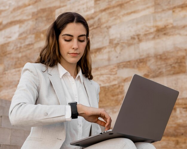 Femme d'affaires élégante regardant smartwatch et travaillant sur un ordinateur portable à l'extérieur