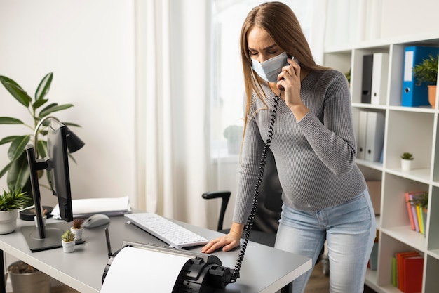 Femme d'affaires enceinte avec masque médical répondant aux appels au bureau