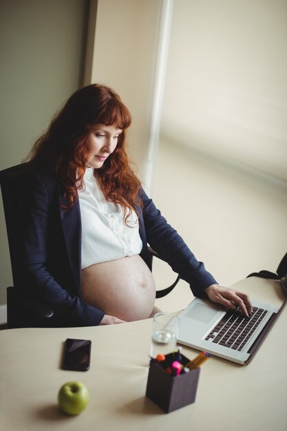 Femme d'affaires enceinte touchant son ventre tout en utilisant un ordinateur portable