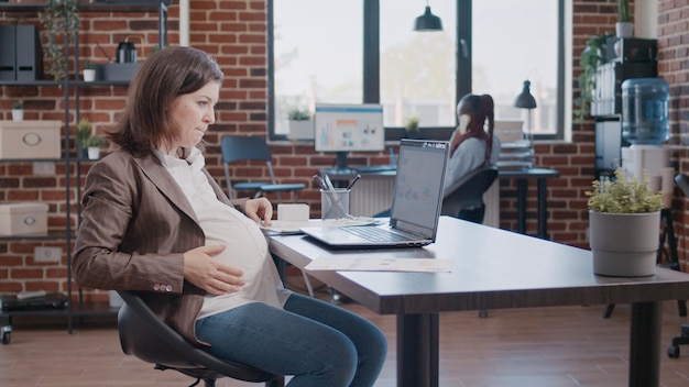 Femme d'affaires enceinte travaillant sur un ordinateur portable à l'aide de documents pour la stratégie marketing dans le bureau de démarrage. Employé attendant un enfant et travaillant sur la planification de projet avec un ordinateur et des fichiers.