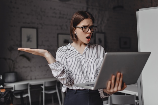 Femme d'affaires étonnée dans des lunettes et des écouteurs tenant un ordinateur portable ouvert et parlant avec indignation sur vidéo dans son bureau.