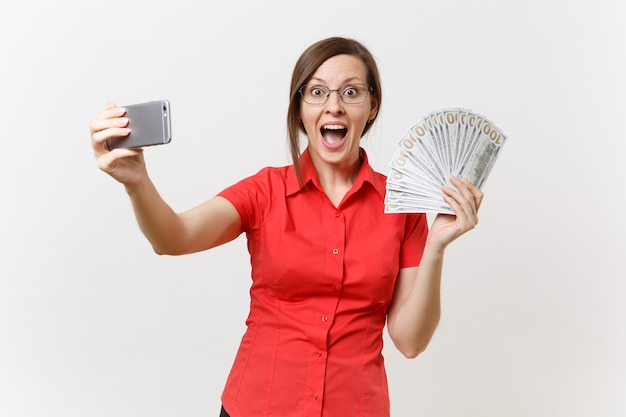 Femme d'affaires excitée en chemise rouge faisant des selfies sur téléphone portable avec beaucoup de dollars, argent liquide isolé sur fond blanc. enseignement de l'éducation dans le concept d'université de lycée.