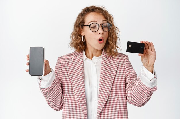 Femme d'affaires excitée dans des verres, montre l'écran du téléphone portable, regarde la carte de crédit avec une expression de visage étonnée sur blanc