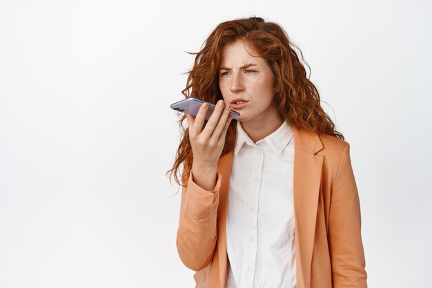 Femme d'affaires frustrée enregistrant un message vocal traduisant sur une application de téléphone mobile avec une expression de visage confus debout sur fond blanc