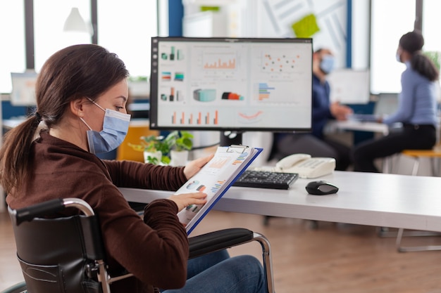 Femme d'affaires handicapée avec masque de protection travaillant dans une nouvelle société financière commerciale normale tapant sur un ordinateur, vérifiant les rapports