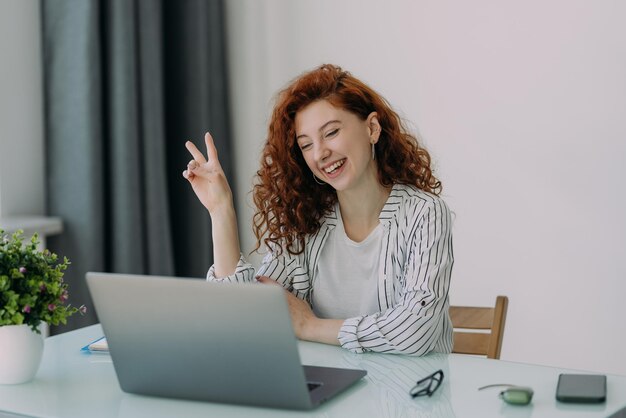 Une femme d'affaires indépendante aux cheveux roux est assise devant un ordinateur portable communique avec des collègues par vidéoconférence est assise au bureau boit du café a une expression heureuse Travail à distance