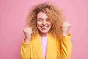 Photo gratuite une femme d'affaires joyeuse et ravie aux cheveux bouclés serre les poings comme un champion célèbre la victoire et entend d'excellentes nouvelles vêtues d'une veste jaune isolée sur un mur rose. femme d'entreprise triomphante