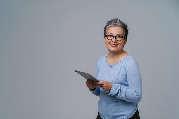 Femme d'affaires en lunettes et tablette numérique dans les mains travaillant en ligne sourire regardant la caméra Jolie femme dans les années 50 en blouse bleue isolée sur blanc Personnes âgées et technologies