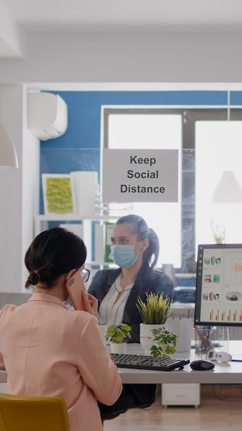 Photo gratuite femme d'affaires avec un masque médical parlant sur un smartphone avec des partenaires pendant que ses collègues travaillent...