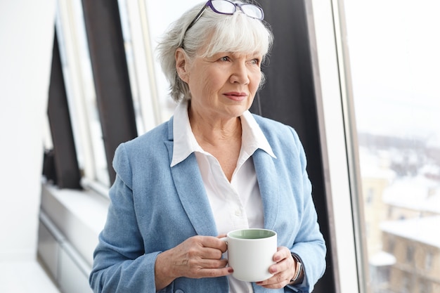 Femme d'affaires mature aux cheveux gris sérieux portant des lunettes sur sa tête et des vêtements formels élégants bénéficiant d'un café chaud, debout près de la fenêtre avec une tasse dans ses mains, ayant un regard pensif pensif