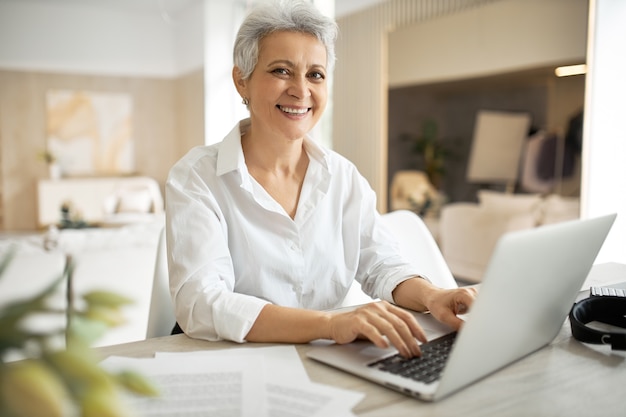 Femme d'affaires mature élégante avec coupe de cheveux courte assis devant un ordinateur portable, regardant l'écran avec la bouche ouverte comme si elle disait quelque chose
