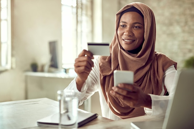 Femme d'affaires musulmane souriante faisant des achats en ligne tout en utilisant une carte de crédit et un téléphone portable au bureau