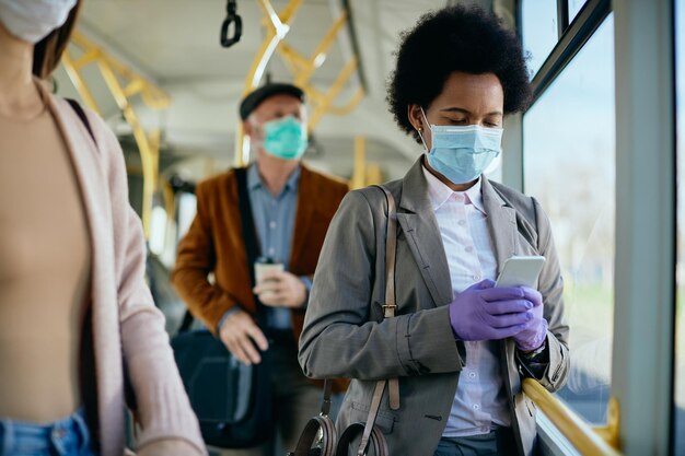 Femme d'affaires noire envoyant des SMS sur son téléphone portable tout en portant un masque de protection et des gants dans les transports en commun