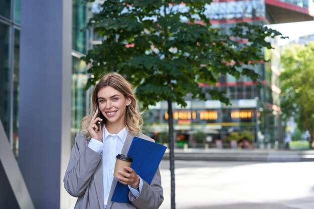 Une femme d'affaires occupée boit des pourparlers de café sur un téléphone portable détient des documents pour des promenades au travail dans le centre-ville