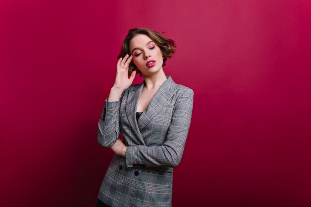 Femme d'affaires pensif avec maquillage rose posant sur un mur bordeaux. Sérieuse jeune femme en veste grise avec des cheveux courts brillants pensant à quelque chose.