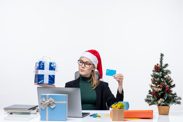 Femme d'affaires positive avec chapeau de père Noël et portant des lunettes assis à une table tenant un cadeau de Noël et une carte bancaire sur fond blanc