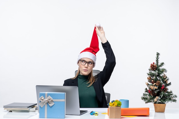 Femme d'affaires positive jouant avec son chapeau de père Noël assis à une table avec un arbre de Noël et un cadeau dessus et penser à quelque chose sur fond blanc