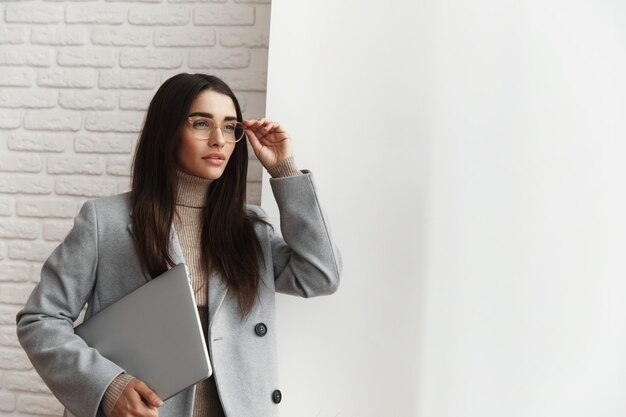 Femme d'affaires professionnelle dans des verres debout près de la fenêtre avec un ordinateur portable.