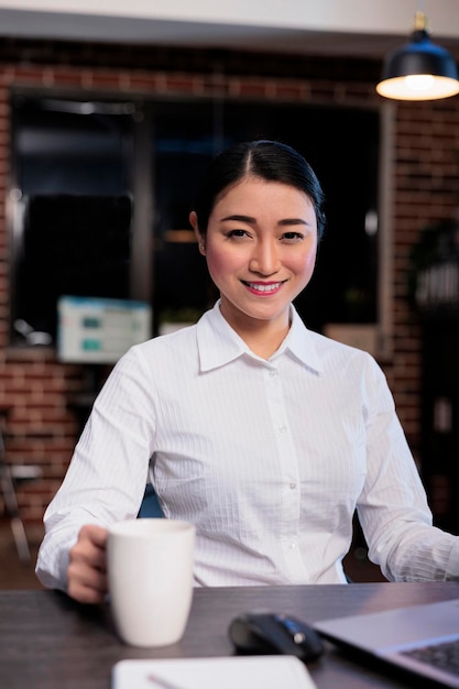 Femme d'affaires à la recherche confiante assise au bureau dans l'espace de travail de bureau tout en souriant à la caméra. Employé de la start-up sur le lieu de travail assis au bureau de l'ordinateur tout en regardant la caméra.