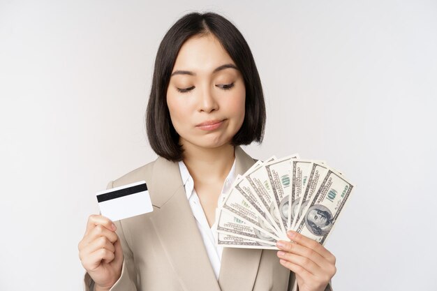 Femme d'affaires réfléchie femme d'entreprise coréenne montrant une carte de crédit et de l'argent en dollars en mains debout sur fond blanc et pensant