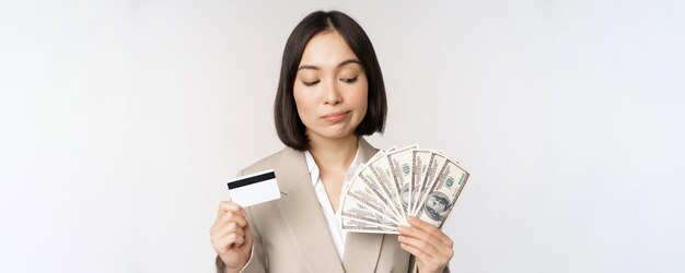 Femme d'affaires réfléchie femme d'entreprise coréenne montrant une carte de crédit et de l'argent en dollars en mains debout sur fond blanc et pensant