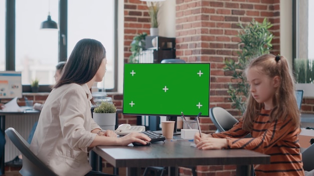 Femme d'affaires regardant un écran vert pendant que sa fille dessine sur papier au bureau du lieu de travail. Mère qui travaille à l'aide d'un modèle isolé et d'un arrière-plan de maquette avec clé chroma, ayant un enfant au travail