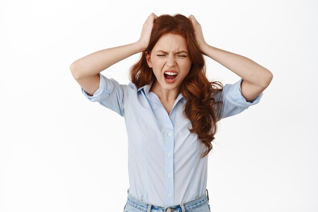 Une femme d'affaires rousse frustrée crie de tension et de détresse, tient les mains sur la tête troublée, a un gros problème, perd, debout sur fond blanc