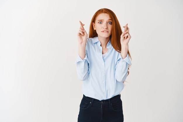 Femme d'affaires rousse pleine d'espoir priant croiser les doigts pour la bonne chance et regardant la caméra pour les résultats fond blanc