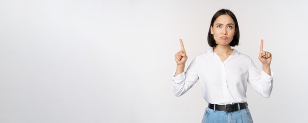 Femme d'affaires sceptique chef de bureau asiatique pointant les doigts vers le haut et grimaçant douteux hésitant debout sur fond blanc