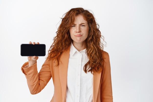 Une femme d'affaires sceptique montre un écran de téléphone horizontal et boude un smartphone déçu qui n'aime pas debout sur fond blanc