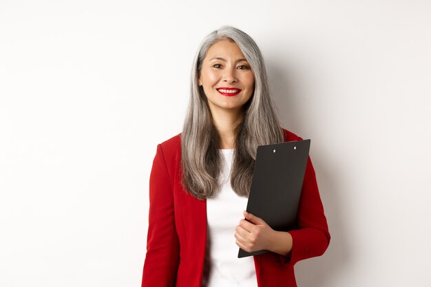 Femme d'affaires senior asiatique réussie tenant un presse-papiers, portant un blazer rouge et du rouge à lèvres au travail, souriant à la caméra, fond blanc