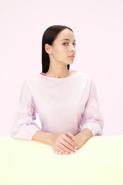 Femme d'affaires sérieuse assise à table sur un fond de studio rose. Le portrait dans un style minimalisme