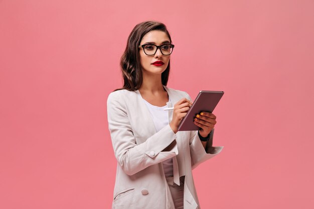 Femme d'affaires sérieuse avec des lèvres rouges vives en tenue élégante beige et des lunettes tient la tablette sur fond isolé rose.